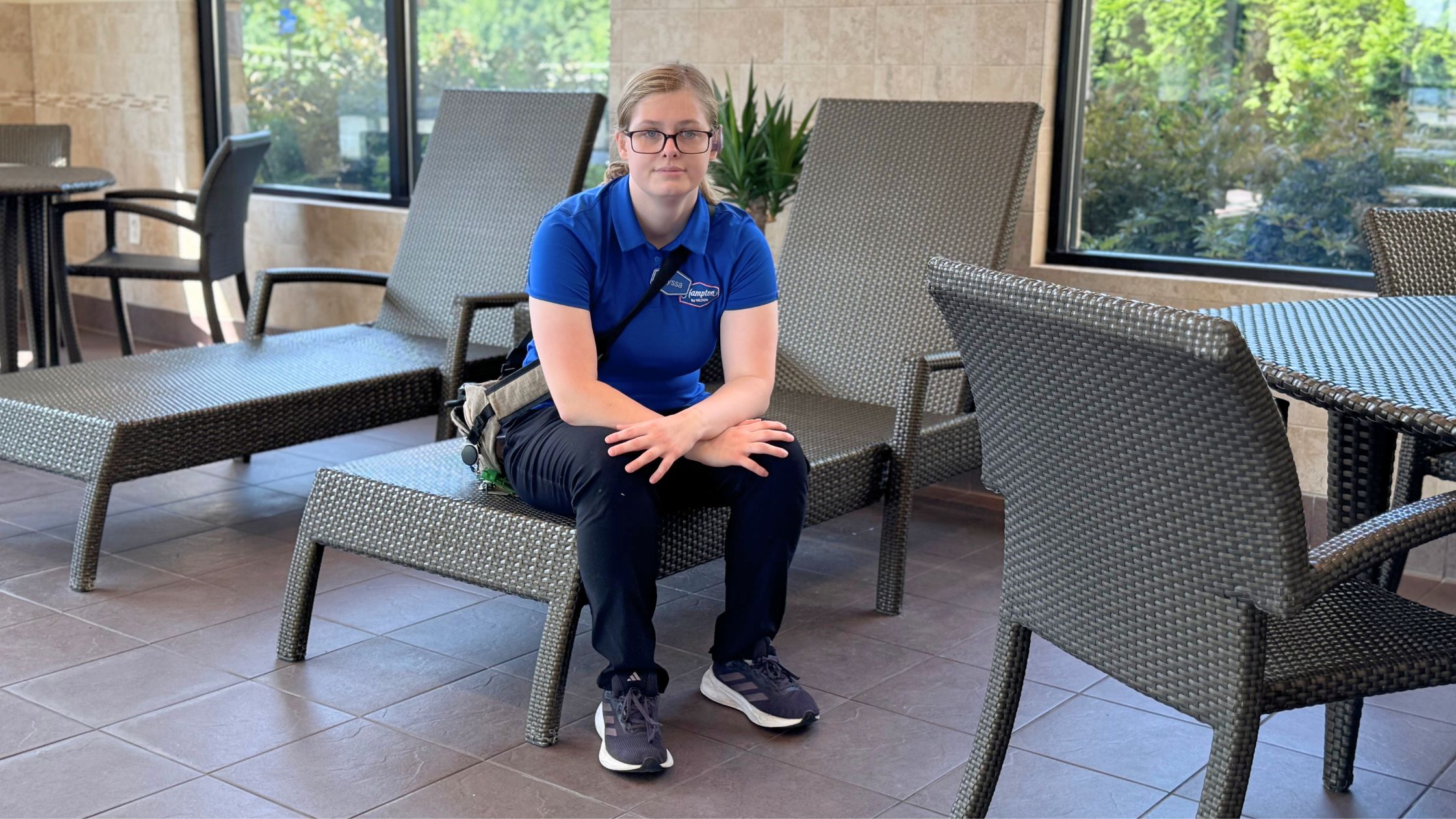 Alyssa sits on a brown woven lounge chair in the hotel indoor pool area wearing a bold blue collared work shirt and black pants and sneakers.
