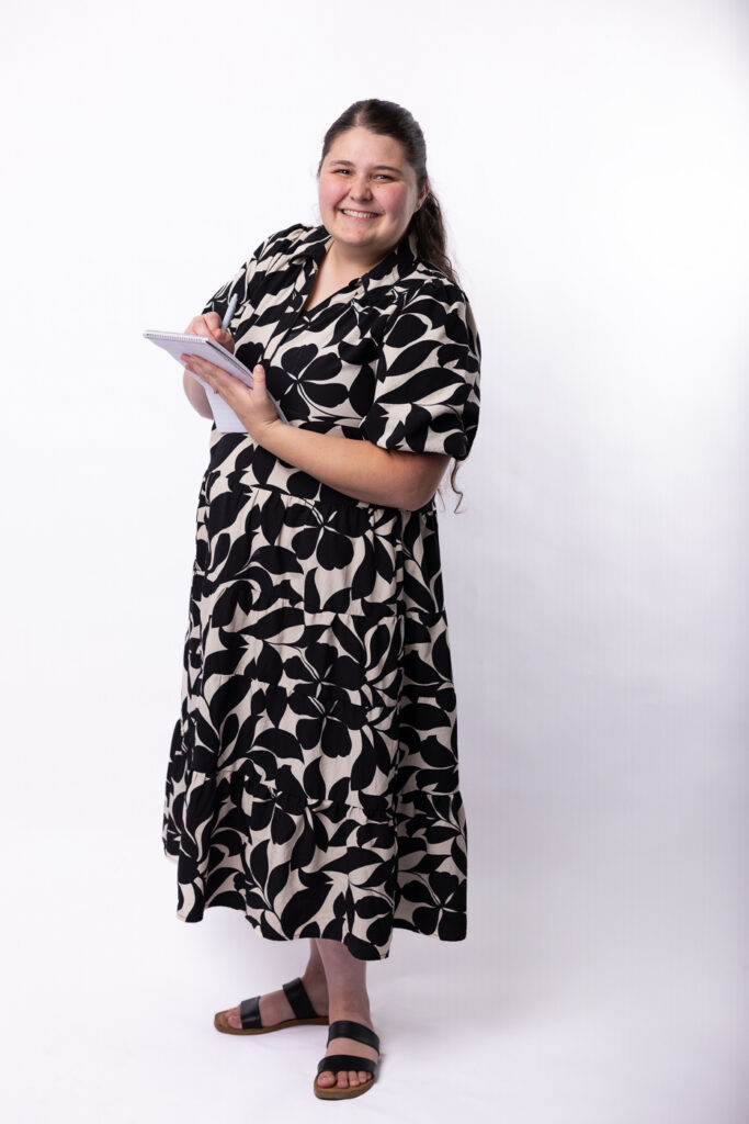 Hannah standing with a note pad and pen in her hands, writing and smiling at the camera.