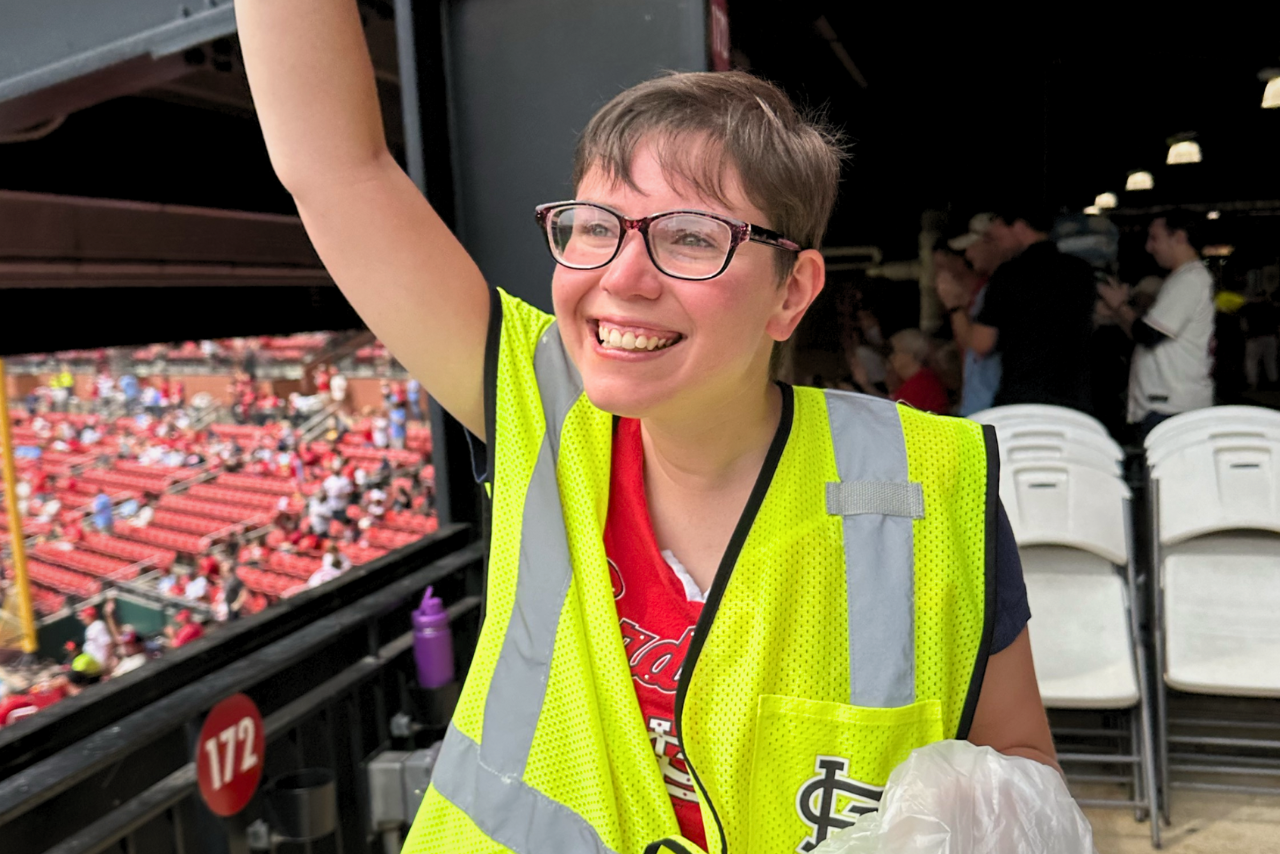 Allison smiles and looks out over the Cardinals baseball stadium, wearing a neon yellow work vest and holding a plastic bag to collect recycleables.