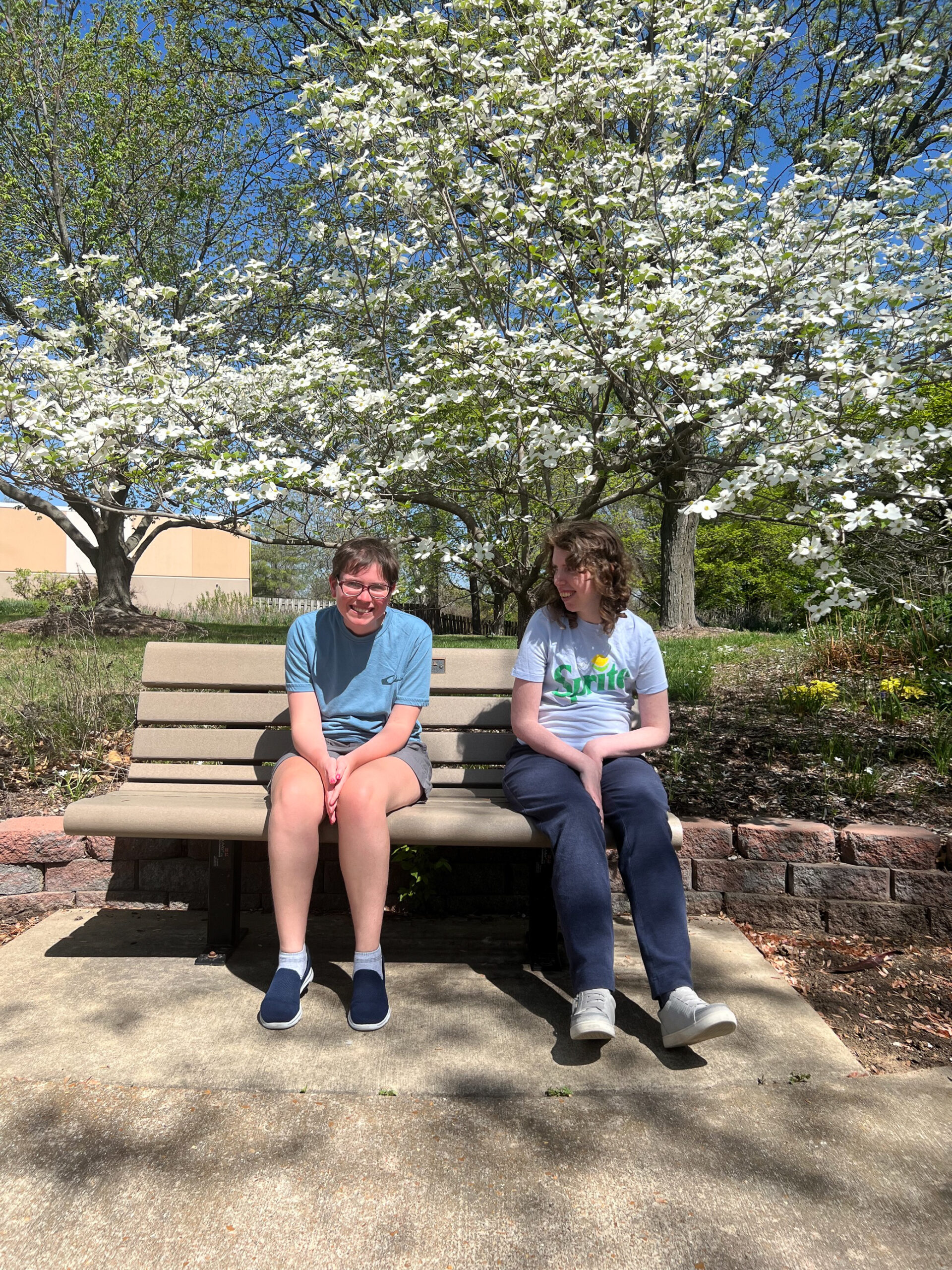 Mahon center participants Allison and Kelsey sit next to eachother on a bench at a park with blooming, flowering trees behind them